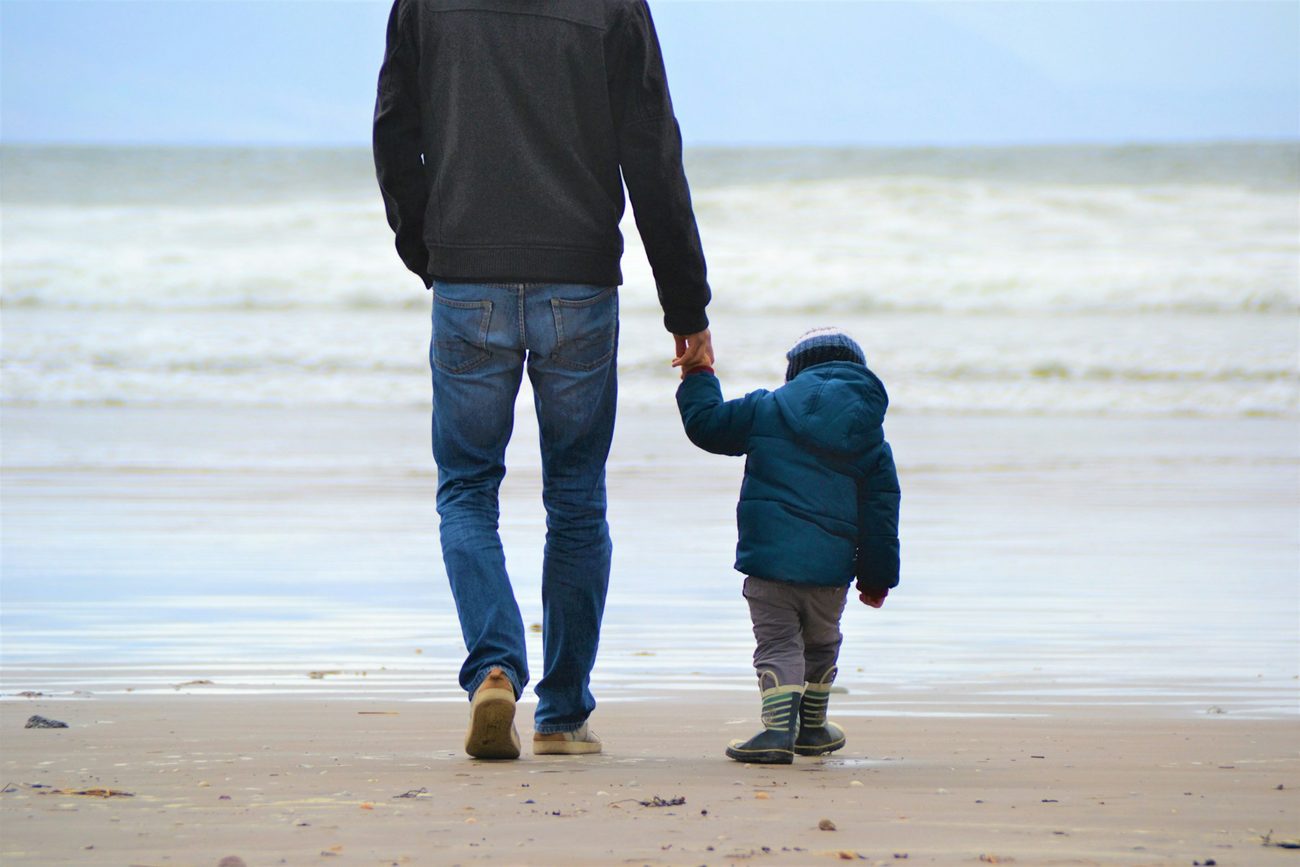 A father walking along the shoreline holding his young son's hand, both seen from behind